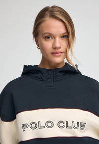 Huvtröja i marinblått med krämfärgad horisontell rand och broderad "POLO CLUB"-logotyp. Har knäppning vid halsen och mjuk textur.