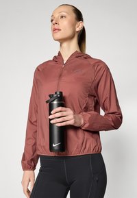 Vandtæt, rustfarvet jakke med hætte, elastiske mansjetter og lynlås foran. Sort sportsflaske med hvidt logo. Sorte leggings.