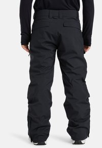 Persoon gekleed in zwarte waterdichte outdoorbroek met achterzakken en ritsen bij de enkel, staand tegen een witte achtergrond.