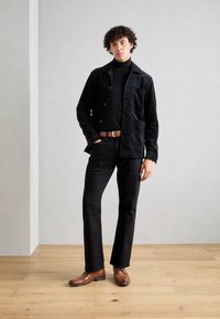 Veste-chemise en velours côtelé noir avec boutons et poche avant, associée à un col roulé noir et un pantalon noir taillé sur mesure. Chaussures en cuir marron.