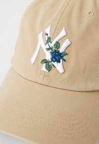 Beige Kappe aus strukturiertem Stoff mit einem weißen gestickten Logo und blauen Blumenakzenten. Gebogener Schirm mit sichtbaren Nahtdetails.