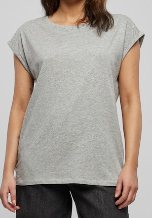 Femme portant un t-shirt gris clair uni à manches courtes retroussées et un pantalon noir sur fond uni.