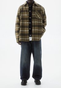 Camisa de franela de cuadros marrones y beige, superpuesta sobre una camisa de mezclilla oscura, combinada con jeans azules holgados y zapatillas negras.