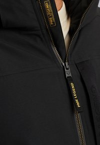 Giubbotto nero caratterizzato da un tessuto texturizzato, con una zip prominente e dettagli in oro, e un'etichetta con il logo. La zip è centrale e funzionale.