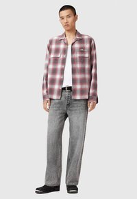 Jonge man in een rood en grijs geruite blouse over een wit tanktop, met grijs gewassen jeans, zwarte schoenen, en staande tegen een effen achtergrond.