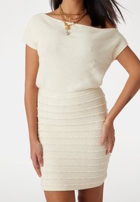 Haut en maille décolleté couleur crème, associé à une mini-jupe moulante texturée avec des rayures horizontales et des accents subtils de perles.