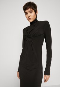 Robe noire à manches longues avec un col montant, un détail en avant torsadé et une silhouette ajustée, fabriquée en tissu lisse et élastique.