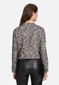 Fekete, fehér és bézs színű, texturált, cropped fazonú dzseki, szőtt mintával, hosszú ujjakkal és rojtos aljjal. Fekete nadrággal párosítva.