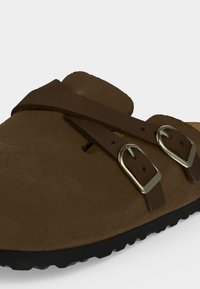 Καφέ δερμάτινο σανδάλι τύπου slip-on με δύο ρυθμιζόμενα λουράκια και ασημένιες μεταλλικές αγκράφες, με υφασμένη μαύρη σόλα.