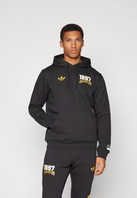 Svart hoodie med känguruficka, prydd med gul "1897 JUVENTUS"-text och Adidas-logotyp. Tillverkad av mjukt material med en avslappnad passform.