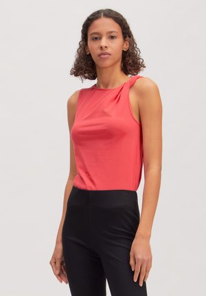 Vrouw met krullend haar die een mouwloze koraalkleurige top en hooggetailleerde zwarte broek draagt, staand tegen een effen lichte achtergrond.