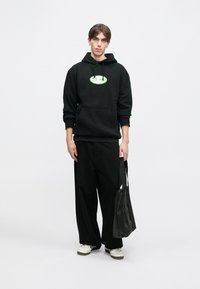 Hanorac negru cu grafică verde, cu un buzunar frontal, asortat cu pantaloni negri largi. Modelul ține o geantă de umăr neagră și poartă adidași albi.