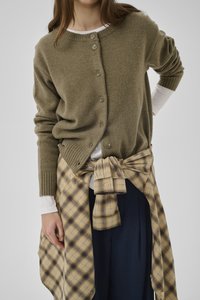 Frau trägt eine olivgrüne, geknöpfte Strickjacke über einem weißen Langarmshirt, mit einem gelb-karierte Hemd um die Taille gebunden und marineblaue Hose.