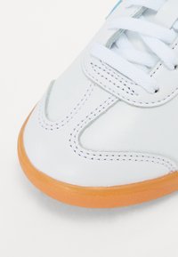 Sapatilha de couro branco com uma superfície lisa, sola em borracha laranja e atacadores brancos. Apresenta costura ao longo da parte superior para um detalhe adicional.