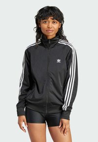 Sort zip-up jakke lavet af glat stof, der har tre hvide striber på hver ærme og et lille hvidt logo på brystet.