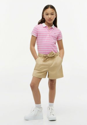 Jeune fille portant un polo rayé rose et blanc, un short beige ceinturé, des chaussettes blanches et des baskets blanches, debout les mains dans les poches.