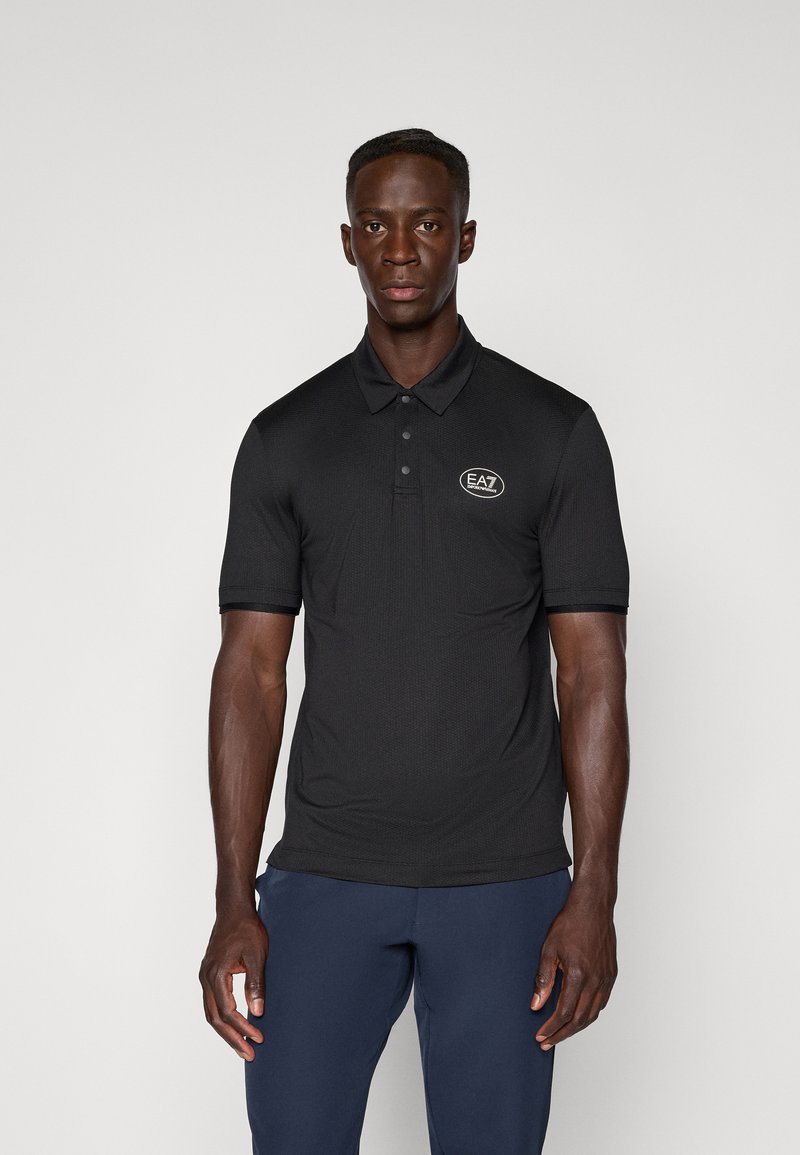 Polo noir avec un col à trois boutons, manches courtes, et logo EA7. Le tissu présente une texture subtile. Associé à un pantalon bleu marine.