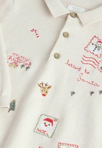 Crèmekleurige gebreide polo met borduursels van kerstmotieven: een rendier, een sneeuwman, een zuurstok en tekst in rood en groen. Drie knopen.