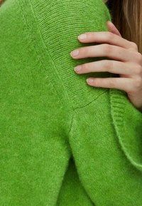Leuchtend grüner Strickpullover mit weicher Textur, ausgestattet mit gerippten Akzenten an Schulter und Ärmel, teilweise gezeigt mit einer Hand, die darauf ruht.