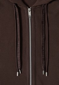 Primo piano di felpa con cappuccio marrone scuro a coste con cerniera argentata e coulisse abbinate con testo in rilievo a ripetizione.