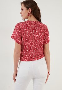 Rød blomstrete bluse med korte ærmer, der har hvide og gule blomstermønstre. Stylet med hvide jeans, der viser et afslappet design.