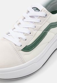 Vans Sneaker mit einem cremefarbenen Canvas-Obermaterial, grünen Lederakzenten, weißen Schnürsenkeln und einer strukturierten Gummisohle mit schwarzer Umrandung.