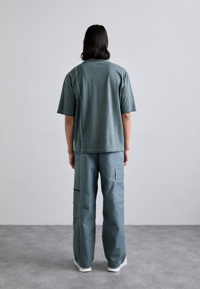 Camiseta gris de gran tamaño con mangas cortas, combinada con pantalones cargo azul claro que presentan bolsillos laterales y detalles con cremallera. Ropa casual.