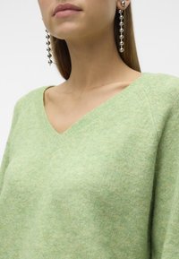 Grüner V-Ausschnitt-Pullover mit weichem Material und entspannter Passform. Lange silberne Ohrringe mit runden Perlen ergänzen das Outfit.