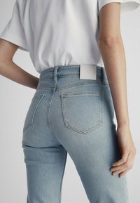 Light blue denim jeans with a high waist, featuring two back pockets and a white leather patch at the waistband. Smooth texture, form-fitting design.
