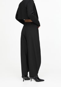 Blazer noir court à manches longues, associé à un pantalon noir à jambes larges et des talons pointus noirs. Texture lisse, coupe ajustée.