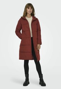 Manteau rembourré rouge avec capuche, affichant une fermeture éclair à l'avant, des poches latérales et un matelassage horizontal. Porté avec un pull crème et un pantalon noir.