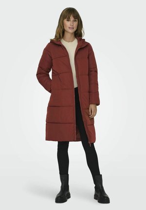 Manteau rembourré rouge avec capuche, affichant une fermeture éclair à l'avant, des poches latérales et un matelassage horizontal. Porté avec un pull crème et un pantalon noir.