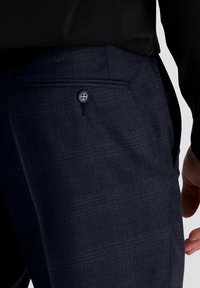 Close-up van donkerblauwe geruite broek met een knoopzak aan de achterkant, gedragen met een zwart bovenstuk en een gedeeltelijk zichtbare hand.