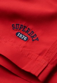 Tissu en coton rouge avec des plis repliés, présentant une broderie "SUPERDRY 1976" en bleu marine. Design simple et texture lisse.