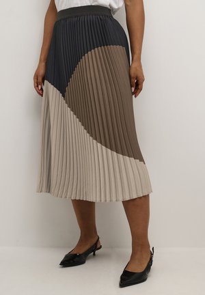 Kobieta nosząca plisowaną spódnicę midi w kolorowych blokach czerni, brązu i beżu, w połączeniu z czarnymi szpiczastymi butami na obcasie typu slingback.