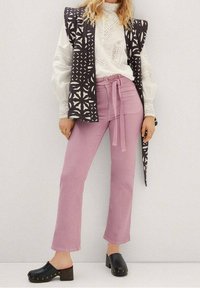 Fioletowe spodnie z wysokim stanem i paskiem do wiązania, połączone z białą bluzką o fakturze oraz kamizelką w czarno-białe wzory. Całość dopełniają czarne klapki.