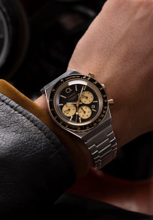 Montre chronographe en acier inoxydable avec une lunette noire et dorée, un cadran noir, des accents dorés et un bracelet en maillons argentés. Elle comporte un tachymètre et un affichage de la date.