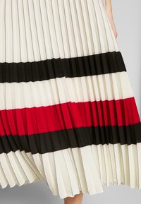Falda con pliegues en tejido crema con distintas franjas horizontales en negro, rojo y negro. Presenta pliegues texturizados y costuras de contraste.