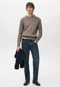 Lichtbruin geribbelde gebreide trui over een wit overhemd, gecombineerd met donkerblauwe jeans en donkerbruine leren laarzen. Houdt een marineblauwe jas vast.