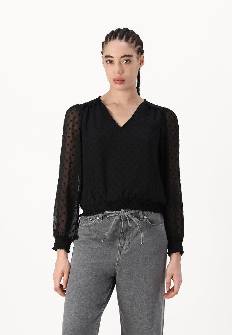 Blouse noire avec des manches transparentes à pois, décolleté en V et taille élastiquée. Associée à un pantalon gris taille haute.