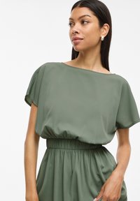 Vestido verde con escote barco, mangas cortas, cintura elástica y textura de tejido fluido. Acabado suave y detalles fruncidos en la cintura.
