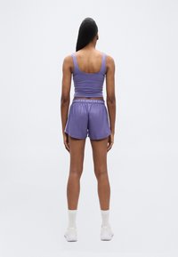 Γυναίκα που στέκεται αντικρίζοντας προς τα πίσω, φορώντας μωβ αθλητική φανέλα και σορτς, λευκές κάλτσες και λευκά αθλητικά παπούτσια, με απλό φόντο.