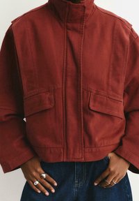 Veste courte rouge foncé en tissu robuste, dotée d'un col haut et de deux grandes poches avant. Associée à un jean en denim bleu.