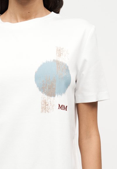 Λευκό βαμβακερό μπλουζάκι με ένα ανοιχτό μπλε κυκλικό γραφικό με υφασμάτινες λεπτομέρειες και κεντημένο «MM» σε κόκκινο χρώμα στο κάτω δεξί μέρος.