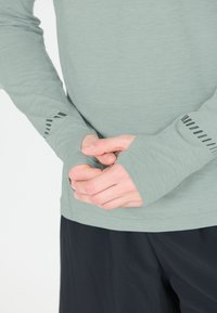 Langdurige mouwen, lichtgroen shirt met duimgaatjes, met een getextureerde afwerking. Zwarte streepaccenten op de manchetten. Gecombineerd met donkere shorts.