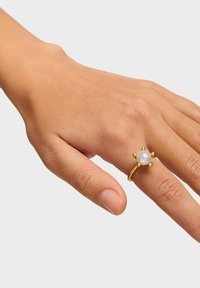 Dłoń z złotym pierścionkiem z pojedynczą białą perłą otoczoną sześcioma małymi złotymi kulkami na palcu serdecznym, na jednolitym tle.