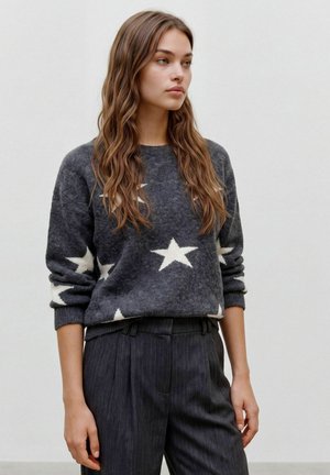Jeune femme aux longs cheveux ondulés portant un pull gris foncé avec des étoiles blanches et un pantalon gris foncé à fines rayures, debout devant un fond uni.