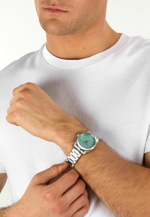 Uomo che regola un orologio da polso argentato con quadrante verde, indossando una t-shirt bianca semplice con scollo rotondo.