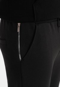 Schwarze Jogginghosen aus weichem Material mit sichtbarem silbernen Reißverschlussdetail an der Seitentasche und einem elastischen Bündchen für einen sicheren Sitz.