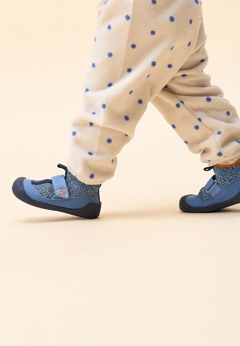 Niebieskie i szare buty z elastycznymi paskami i zapięciem na rzep, z teksturowaną dzianiną, gumową podeszwą, noszone z kremowymi spodniami z kropkami z polaru.
